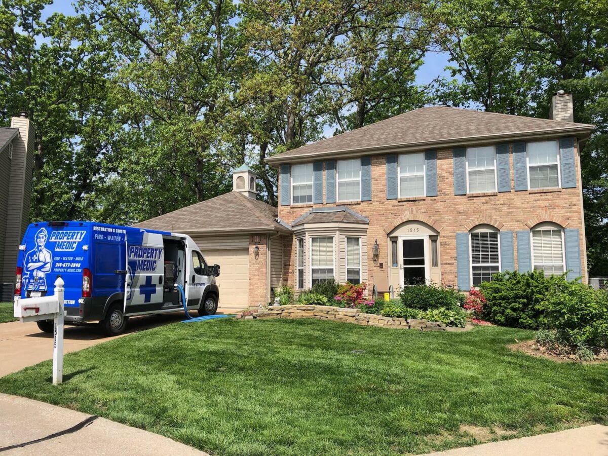 A Property Medics home repair and restoration service van is parked in the driveway of a two-story brick house with green lawn and landscaping.