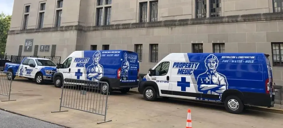 Three "Property Medic" home repair and restoration service vehicles are parked on a street beside a large building, separated from the sidewalk by metal barricades.