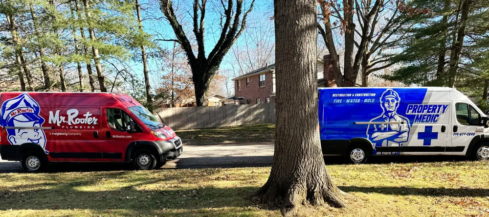 A red Mr. Rooter Plumbing van and a blue Property Medic home repair and restoration service van are parked on a residential street with trees and houses in the background.