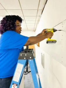 A person standing on a ladder uses a power drill to mount drywall onto a white wall, demonstrating skilled home repair and restoration service inside a building.