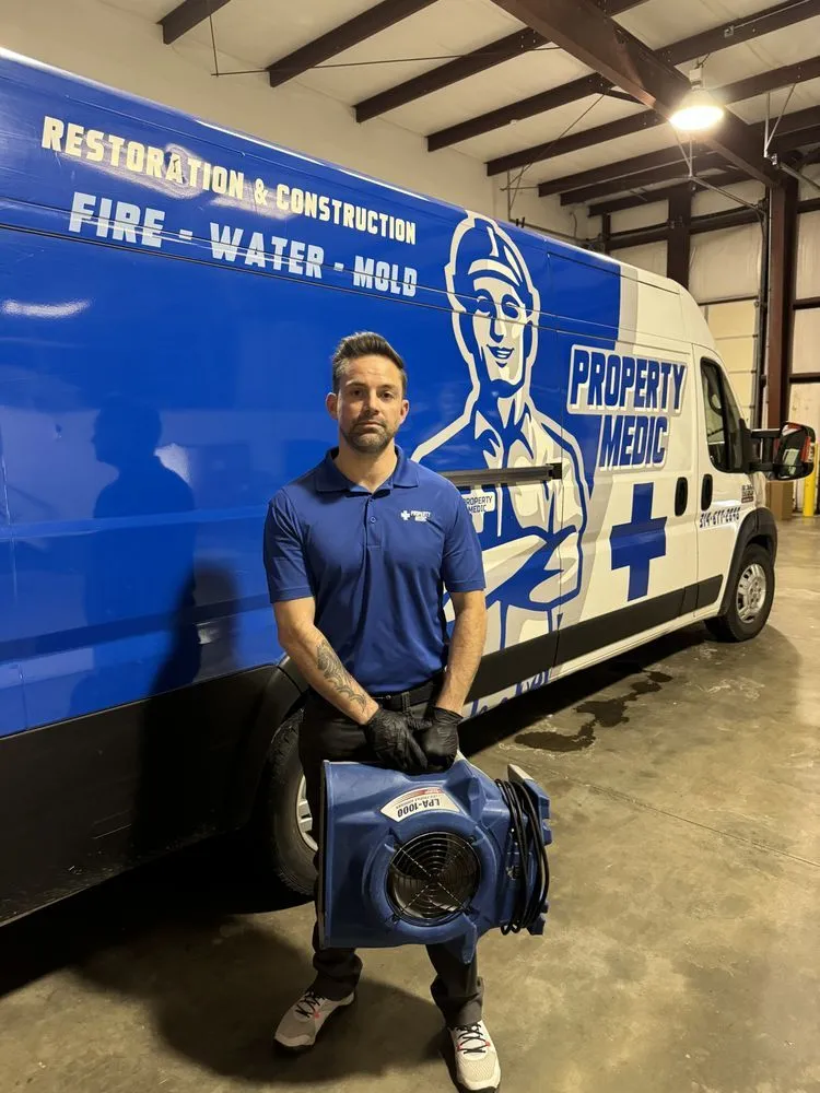 A man in work attire stands in a garage holding an air mover in front of a Property Medic home repair and restoration service van.