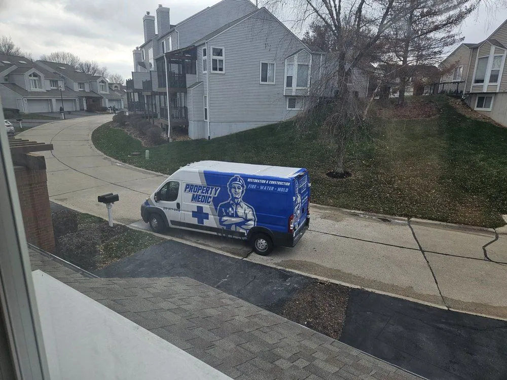 A blue and white van labeled "Property Medic," a trusted home repair and restoration service, is parked on a suburban street in front of townhouses on a cloudy day.